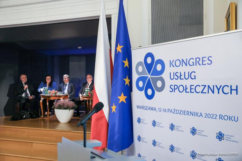 Na pierwszym planie plansza z napisem "Kongres Usług Społecznych. 12-14 października 2022 r." i flagi polski i UE, w oddali kilka osób siedzi na krzesłach i rozmawia