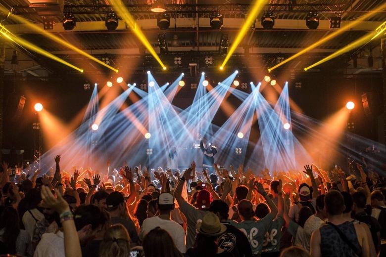 Tłum ludzi na sali koncertowej z wyciągniętymi do góry rękami, scena oświetlona snopami światła, na scenie postać trzymająca mikrofon i śpiewająca