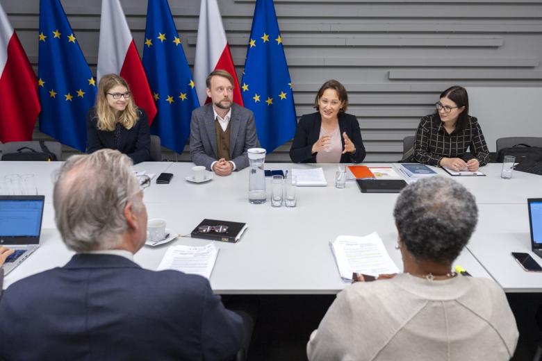 Cztery osoby siedzące przy stole na tle flag Polski i UE