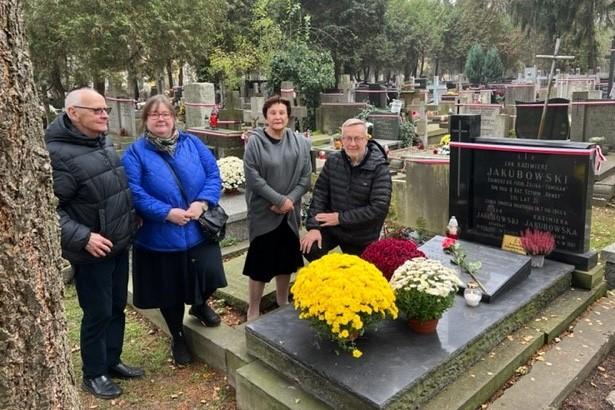 Cztery osoby stoją przy grobie, na którym leżą kwiaty i stoją znicze