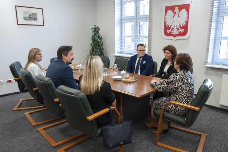 Grupa osób siedzących przy stole, na ścianie za stołem godło Polski