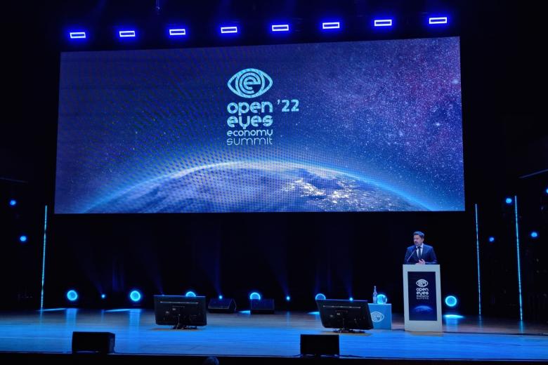 Osoba przemawia na scenie za mównicą na tle dużego telebimu na którym wyświetlana jest grafika ziemi i napis "Open Eyes Economy Summit 2022"