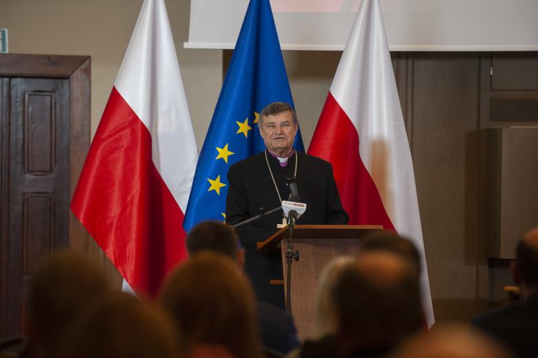Biskup Ryszard Bogusz przemawia na podium na tle flag Polski i Unii Europejskiej. Na dole pierwszego planu widać głowy słuchającej go publiczności.