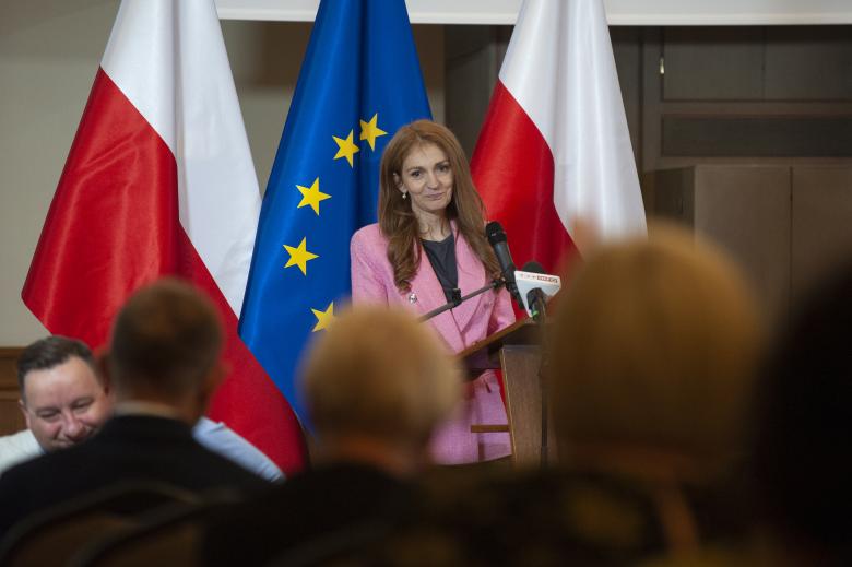 Monika Zima-Parjaszewska przemawia na podium na tle flag Polski i Unii Europejskiej. Na pierwszym planie sylwetki publiczności.