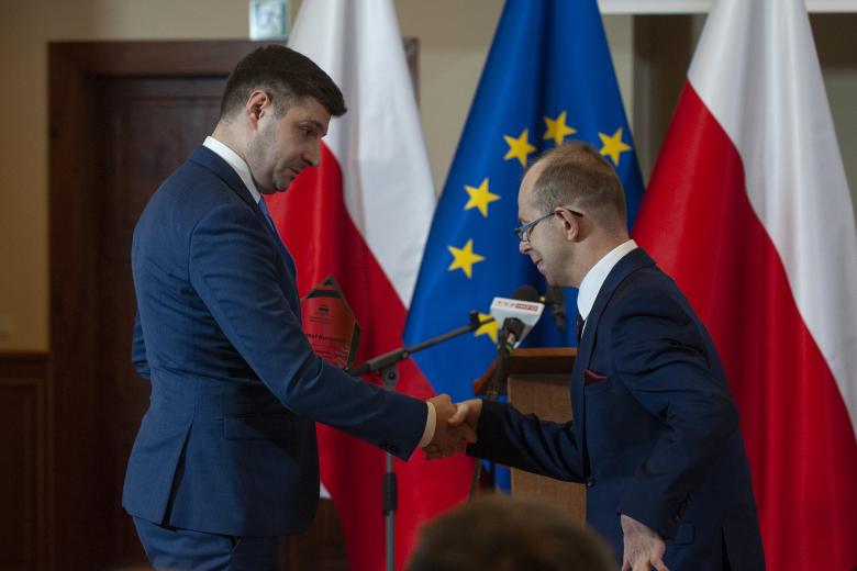 Valeri Vachev wręcza Krzysztofowi Kurowskiemu statuetkę nagrody, ściskając jego dłoń. W tle flagi Polski i Unii Europejskiej.