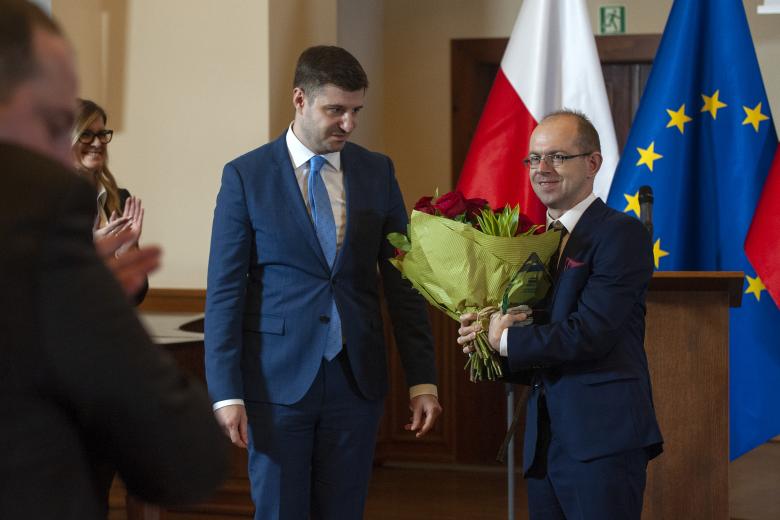 Krzysztof Kurowski stoi uśmiechając się i trzymając kwiaty. Obok niego stoi przyglądający się mu Valeri Vachev. Dookoła uczestnicy uroczystości biją brawo. W tle podium oraz flagi Polski i Unii Europejskiej.