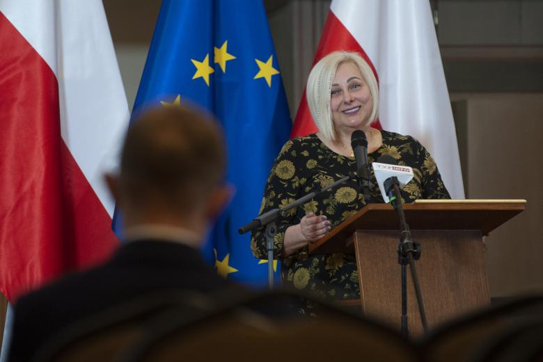 Ewa Pawłowska wypowiada się w stronę laureata, którego tył sylwetki jest na pierwszym planie. W tle flagi Polski i Unii Europejskiej.