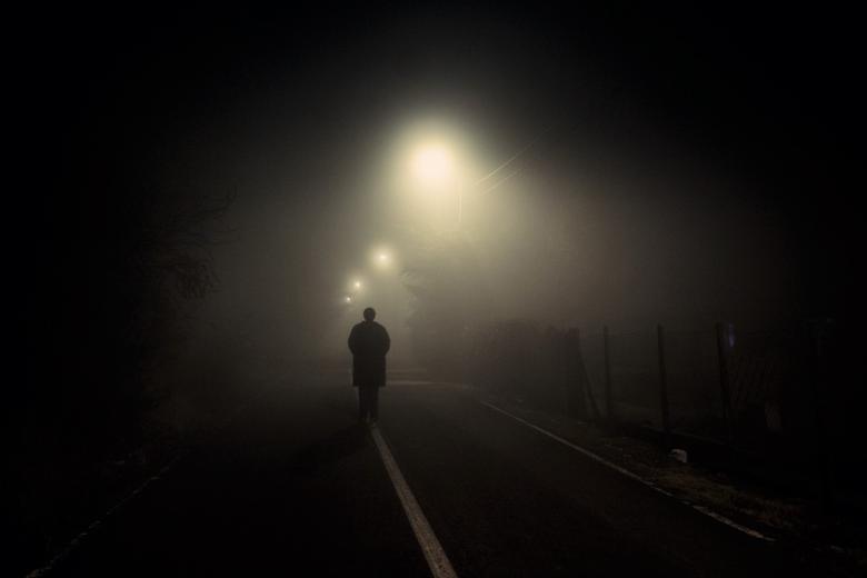 Ciemna postać idąca mglistą nocą środkiem słabo oświetlonej przez latarnie uliczne drogi