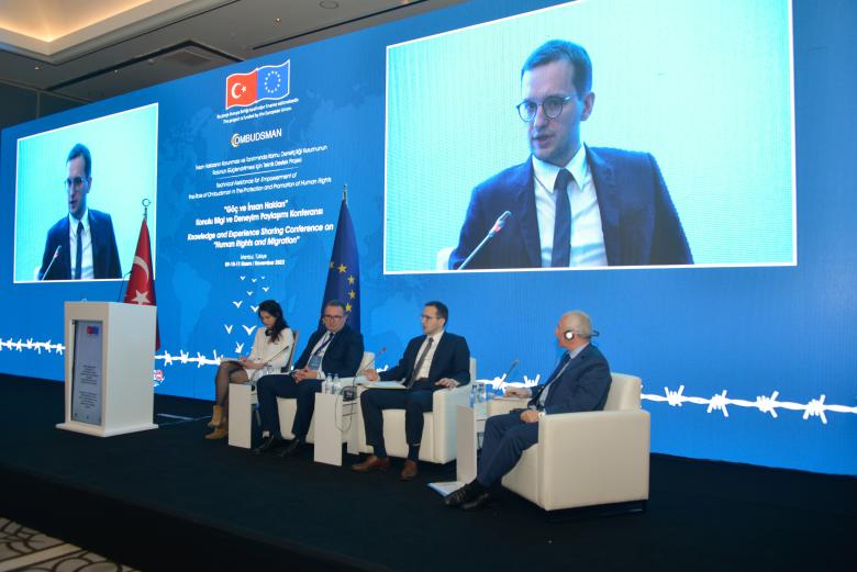 Cztery osoby - jedna kobieta i trzech mężczyzn - siedzą w białych fotelach na scenie. W tle duży ekran przedstawiający temat konferencji i wypowiadającą się osobę oraz flagi Turcji i Unii Europejskiej