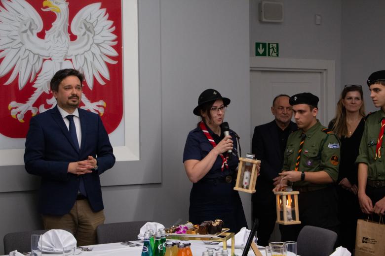 Kobieta w harcerskim mundurze wypowiada się trzymając mikrofon. Po jej prawej stronie stoi RPO Marcin Wiącek, po lewej stronie stoją harcerze i pracownicy biura RPO. W tle godło Polski.