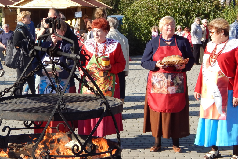 Zdjęcie z lokalnego festynu przedstawiające cztery kobiety w fartuchach i koralach. Jedna z nich trzyma w rękach słoik z przetworami, a druga - bochen chleba. W tle widać przechodzących uczestników festynu i drewniane budki. Na pierwszym planie jest żeliwny grill z rozpalonym drewnem.