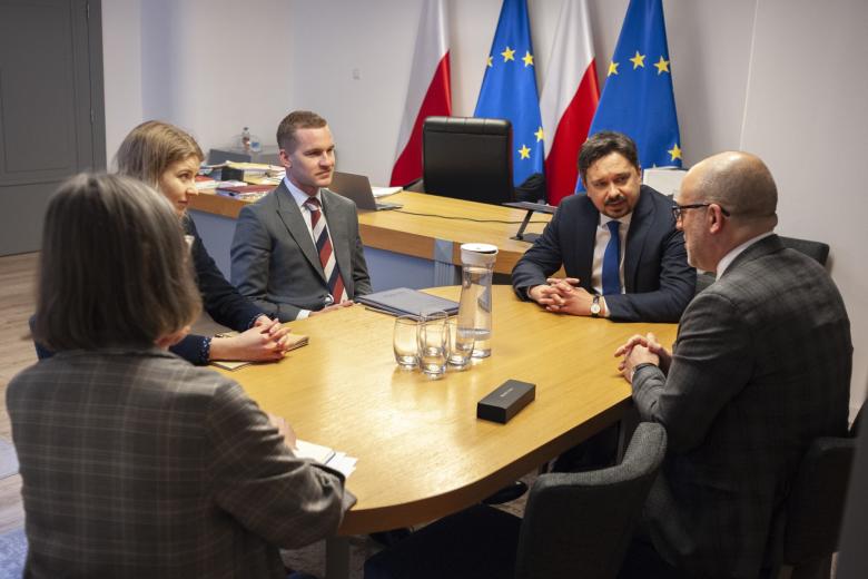 Sześć osób siedzi przy owalnym stole i rozmawia, w tle biurko, za którym stoją flagi Polski i Unii Europejskiej