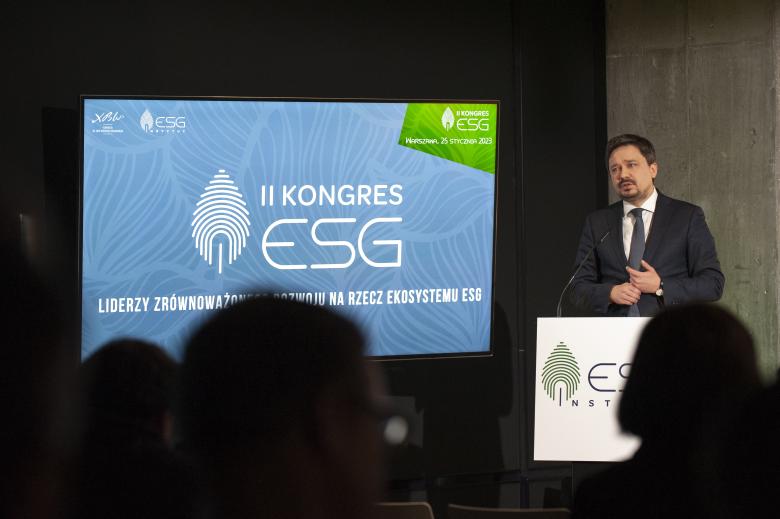 RPO Marcin Wiącek wypowiada się z podium w stronę publiczności. Na ekranie stojącym przy rzeczniku widać logo organizatorów konferencji.
