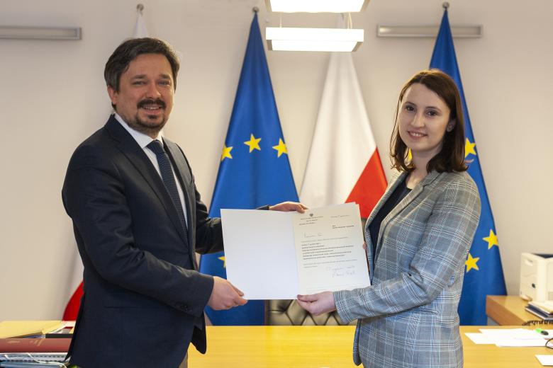 RPO Marcin Wiącek i Monika Wiszyńska-Rakowska uśmiechają się do kamery pokazując dokument powołania na stanowisko. W tle flagi Polski i Unii Europejskiej
