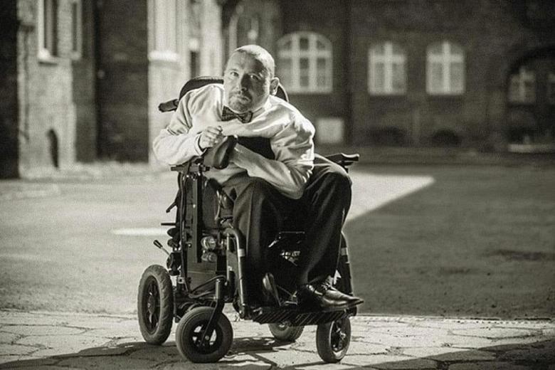 Czarno-białe zdjęcie przedstawiające mężczyznę na wózku inwalidzkim na chodniku, w tle kamienice
