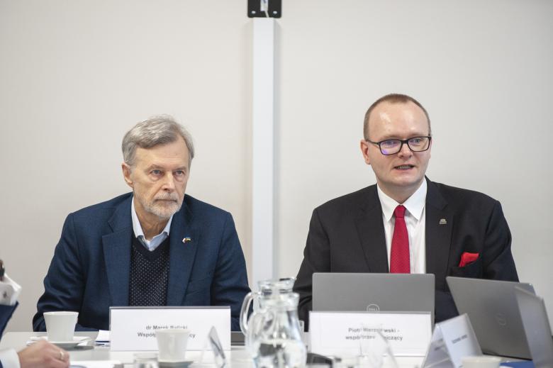 Dwóch współprzewodniczących Komisji: Marek Balicki i wypowiadający się Piotr Mierzejewski siedzą w szczycie konferencyjnego stołu. 