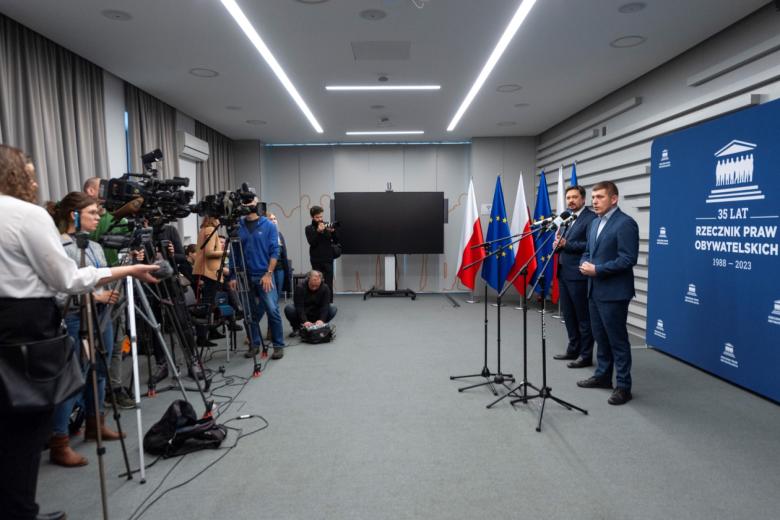 RPO Marcin Wiącek i ZRPO Wojciech Brzozowski stoją przed mikrofonami, po drugiej stronie sali dziennikarze i kilka kamer telewizyjnych