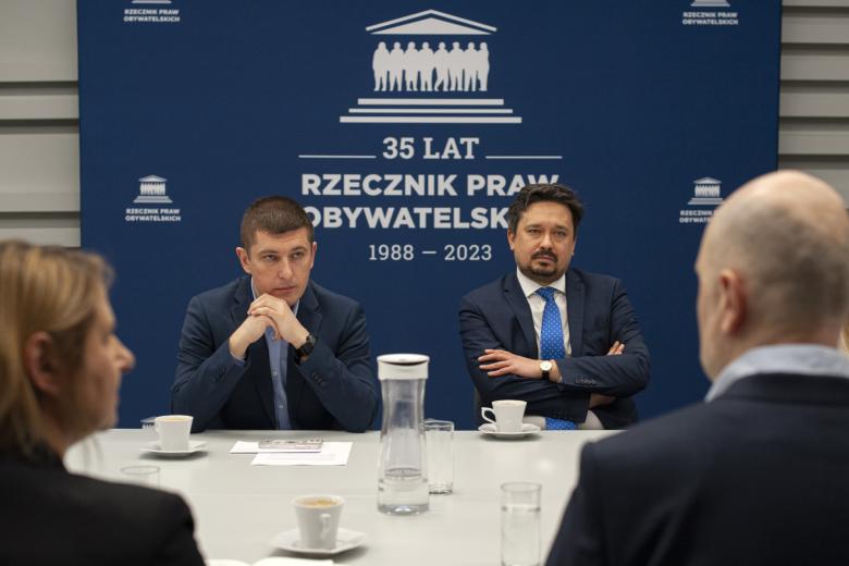 RPO Marcin Wiącek i ZRPO Wojciech Brzozowski siedzą za stołem na tle niebieskiej planszy z napisem "Rzecznik Praw Obywatelskich"