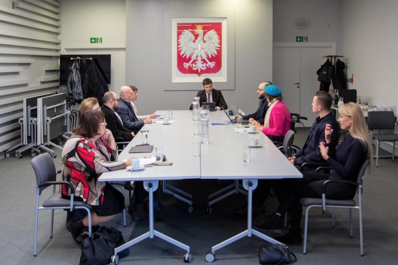 Grupa kilkunastu osób siedzi przy dużym stole konferencyjnym, w tle godło Polski