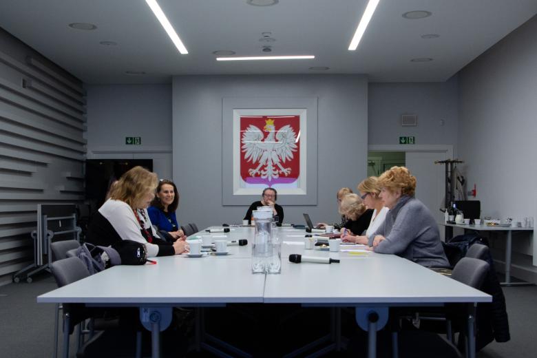 Grupa kilku osób siedzi przy dużym stole konferencyjnym, w tle godło Polski