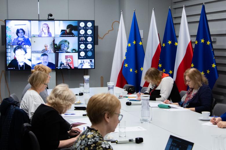 Grupa kilku osób siedząca po obu stronach dużego stołu konferencyjnego, w tle duży ekran z widocznym ekranem z uczestnikami spotkania w aplikacji do komunikowania się na odległość