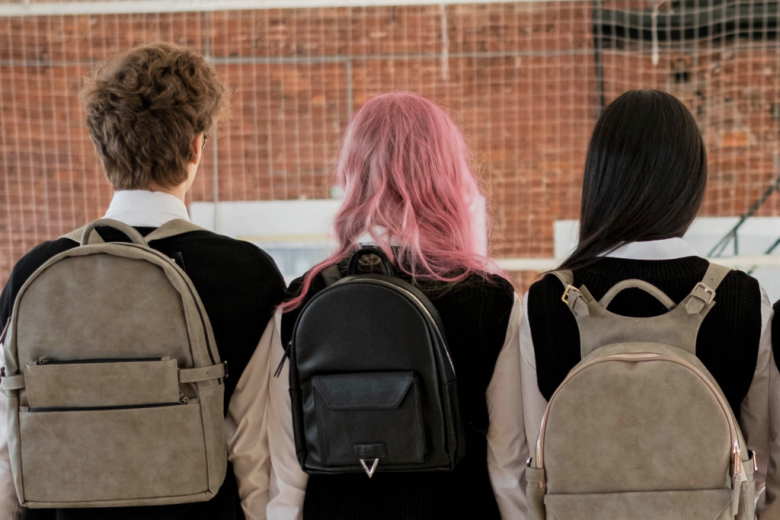 troje uczniów odwróconych plecami o różnych kolorach włosów i z plecakami  