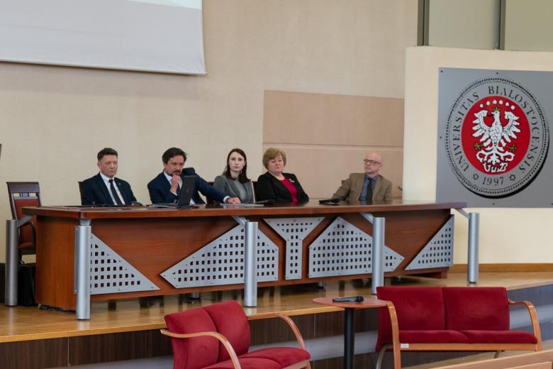 Pięć osób siedzi za stołem na sali wykładowej, z boku grafika orła w koronie z napisem "Uniwersytet w Białymstoku"