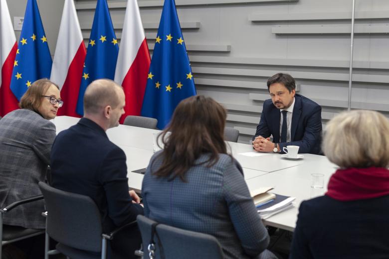 Mężczyzna siedzący za stołem na tle flag Polski i UE słucha osób siedzących po drugiej stronie stołu