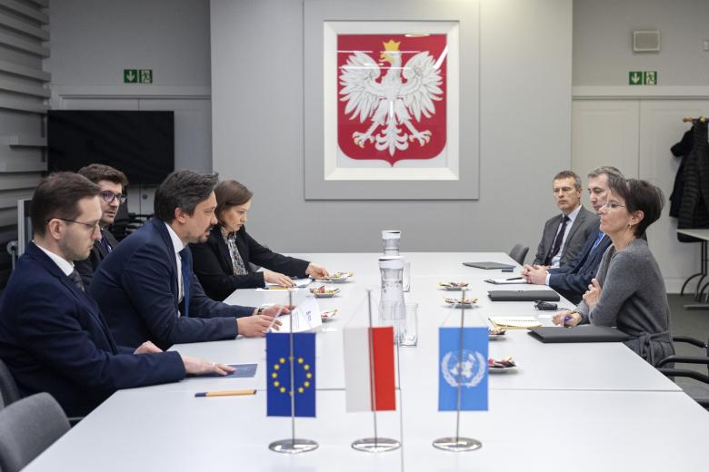 Siedem osób siedzi po obu stronach stołu i rozmawia. W tle na ścianie godło polski, na pierwszym planie małe flagi Polski, UE i ONZ