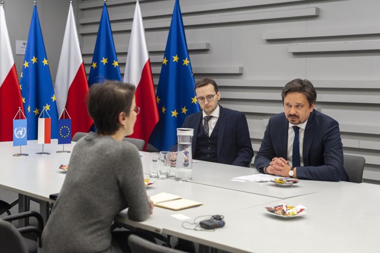Trzy osoby siedzą przy stole i rozmawiają, w tle flagi Polski i UE