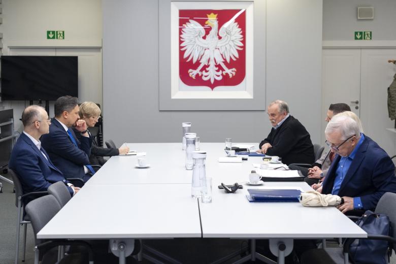 Siedem osób siedzi po obu stronach stołu i rozmawia. W tle na ścianie wiszące godło Polski