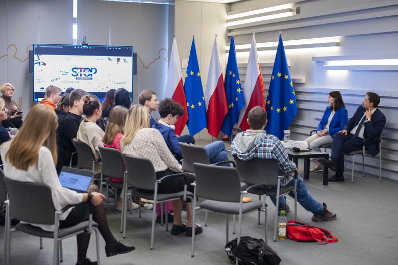 Uczestnicy spotkania oglądają prezentację wyświetlaną na monitorze. W tle flagi Polski i Unii Europejskiej