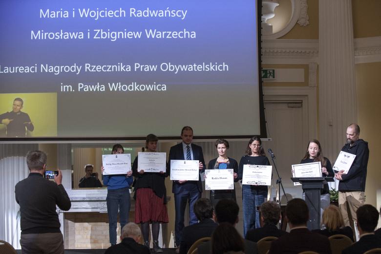 Maria i Zbigniew Radwańscy oraz przedstawiciele Klubu Inteligencji Katolickiej stoją na podium trzymając nekrologi osób zmarłych na granicy Polski i Białorusi