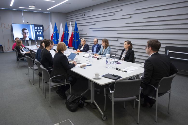 ZRPO Valeri Vachev i członkinie Inicjatywy rozmawiają siedząc przy konferencyjnym stole. W szczycie stołu stoi ekran wyświetlający uczestników spotkania łączących się za pośrednictwem internetu. Obok ekranu stoją flagi Polski i Unii Europejskiej