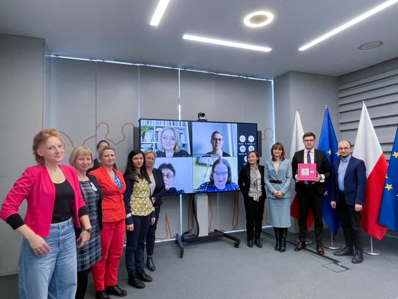 ZRPO Valeri Vachev i uczestniczki spotkania (w tym osoby łączące się za pośrednictwem internetu, które wyświetlane są na ekranie w środku kadru) pozują do zdjęcia. Valeri Vachev trzyma w dłoniach różową skrzynkę. W tle flagi Polski i Unii Europejskiej