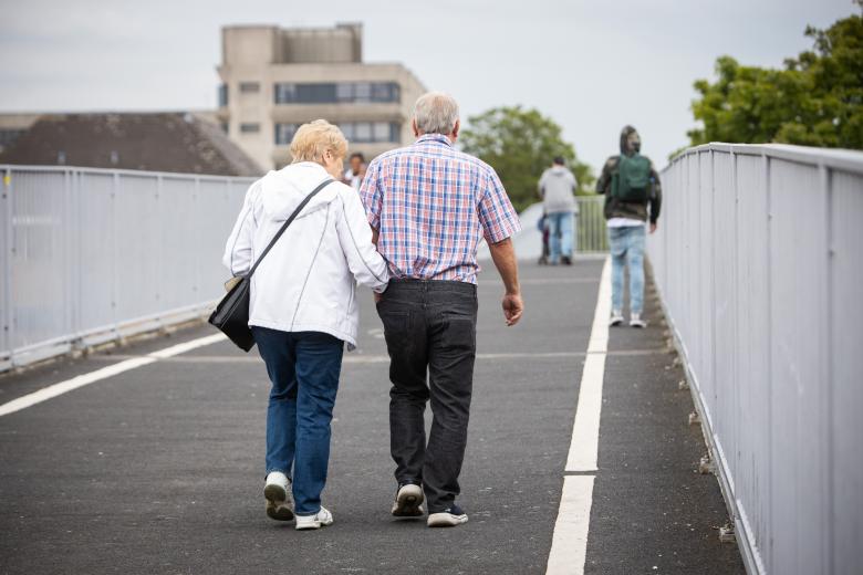 starsza kobieta i mężczyzna idą kładką dla pieszych trzymając się pod rękę.