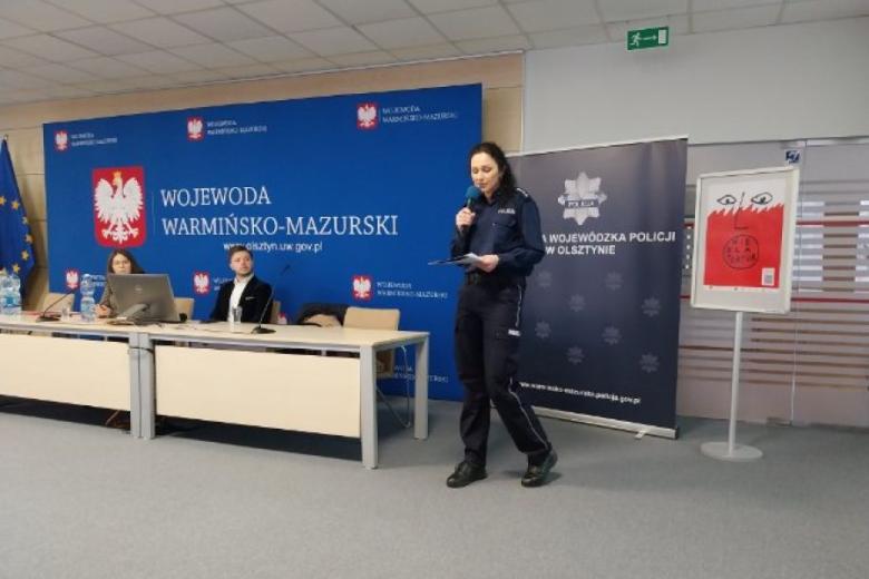 Policjantka w mundurze stoi i mówi do mikrofonu, obok dwie osoby siedzą za stołem, w tle banner z napisem "Komenda Wojewódzka Policji w Olsztynie"