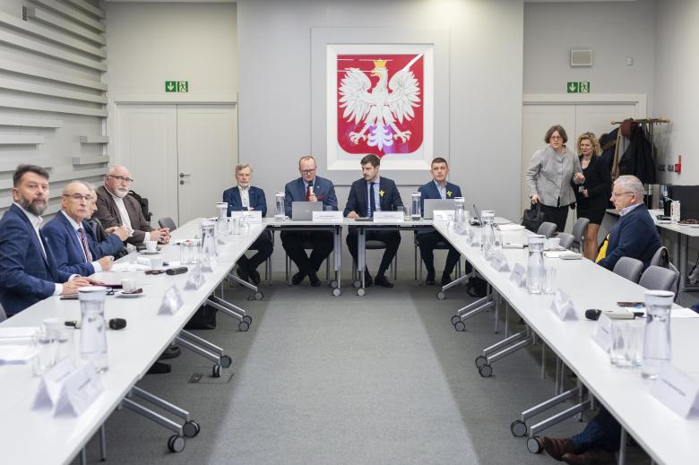 Członkowie Komisji siedzą przy stole. W szczycie stołu siedzi dwóch przewodniczących Komisji oraz zastępcy RPO Valeri Vachev i Wojciech Brzozowski. W tle godło Polski.