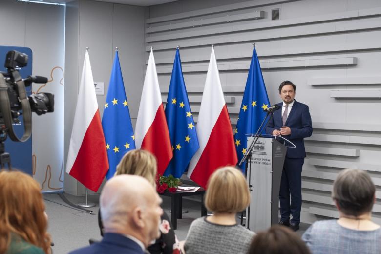 RPO Marcin Wiącek przemawia za mównicą na tle flag Polski i UE