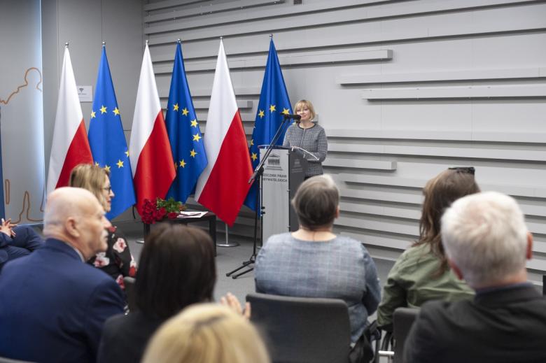 Anna Duniewicz przemawia za mównicą na tle flag Polski i UE