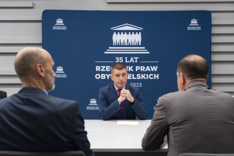 ZRPO Wojciech Brzozowski siedzi za stołem na tle niebieskiej ścianki z napisem "35-lat Rzecznik Praw Obywatelskich" i słucha siedzących na przeciwko osób