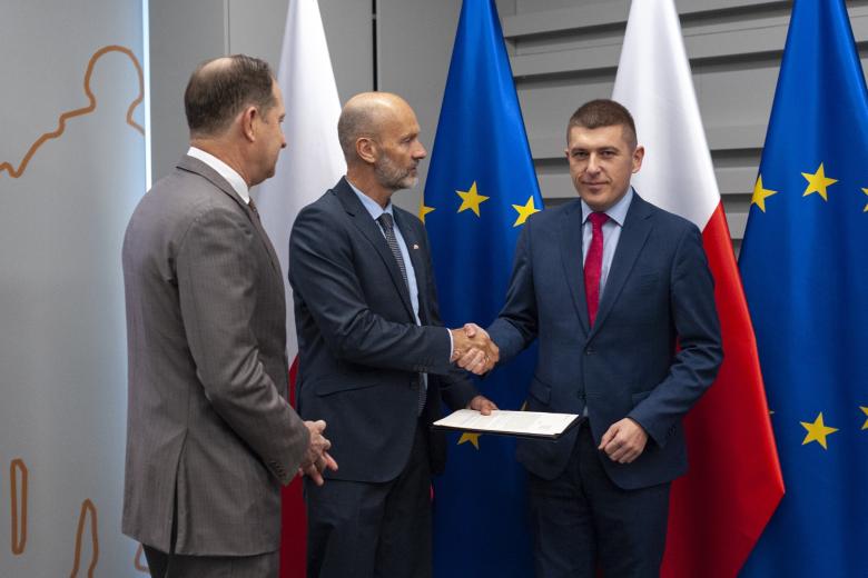 Trzy osoby stoją na tle flag Polski i UE. Środkowa wręcza list ZRPO Wojciechowi Brzozowskiemu z uściskiem dłoni