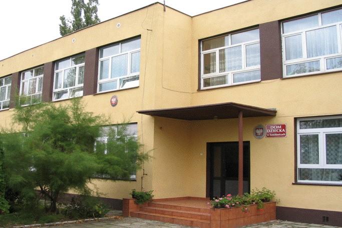 Dwupiętrowy budynek z wejściem, przy którym wisi tabliczka z napisem "Dom dziecka w Tomisławicach"