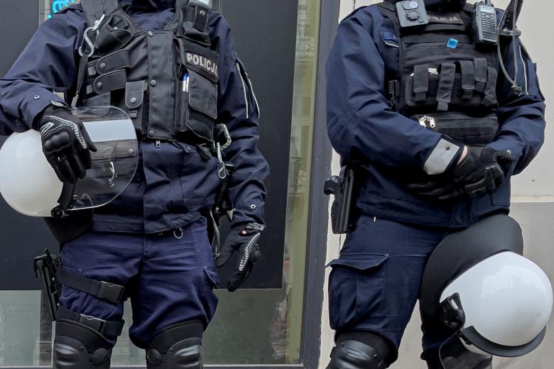 Dwóch policjantów wyposażonych w kamizelki, ochraniacze, hełmy, sprzęt do komunikacji oraz broń palną i białą