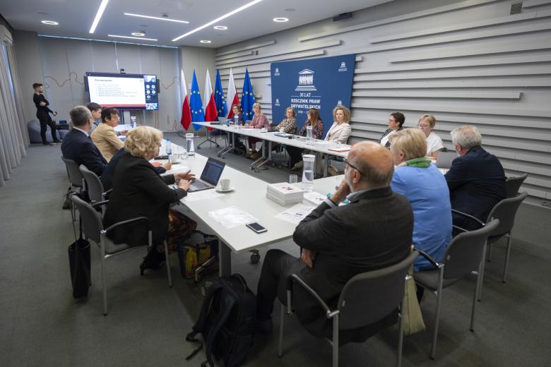 Kilkanaście osób siedzi przy stole konferencyjnym i rozmawia, w tle flagi Polski i UE i duży telewizor z ekranem prezentacji