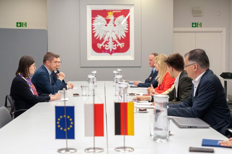 Grupa osób siedzi po dwóch stronach dużego prostokątnego stołu, w tle godło Polski, na pierwszym planie na stole stoją małe flagi Polski, UE i Niemiec