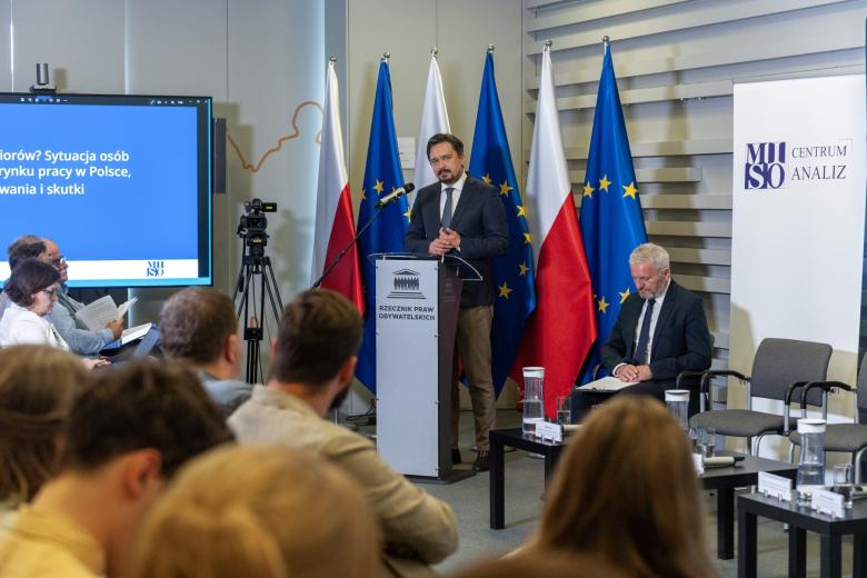 RPO Marcin Wiącek przemawia za mównicą na tle flag Polski i UE