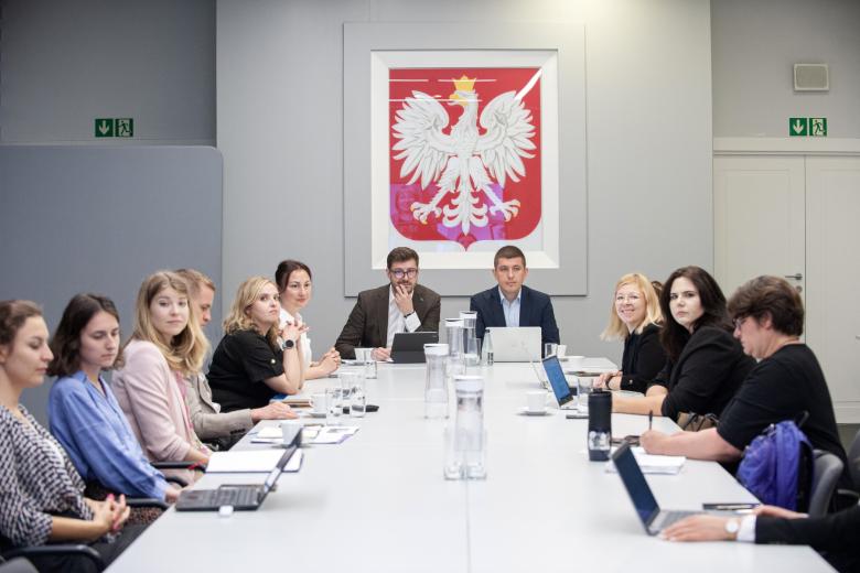Kilkanaście osób siedzi przy dużym stole konferencyjnym, w tle na ścianie godło Polski