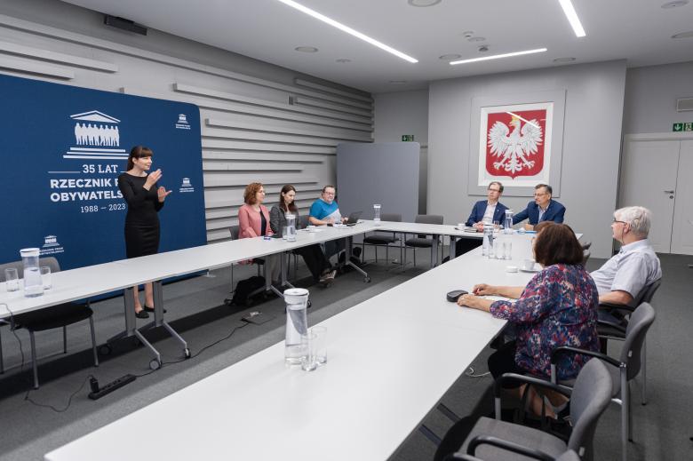 Członkowie Komisji siedzący przy konferencyjnym stole. Obok stołu stoi gestykulująca tłumaczka polskiego języka migowego W tle godło Polski