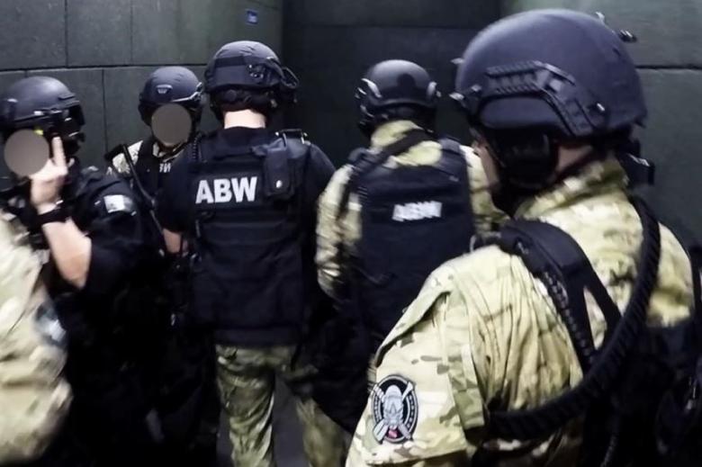 Grupa funkcjonariuszy w kamizelkach z napisem "ABW" na plecach, hełmach i z bronią stoi w korytarzu
